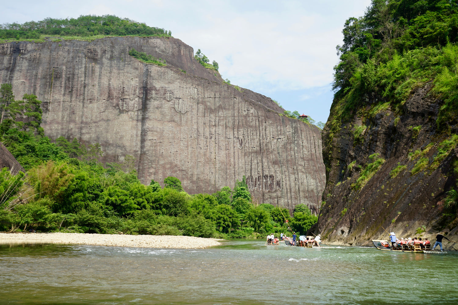 an immense rock cliff behind a river with boats in Wuyi, China.